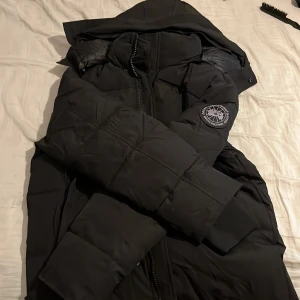 Svart dunjacka från Canada Goose  - Säljer en svart dunjacka från Canada Goose med klassisk patch på ärmen. Jackan har huva, dragkedja och quiltad design som håller dig varm under kalla dagar. Perfekt för dig som vill ha en stilren och funktionell vinterjacka.