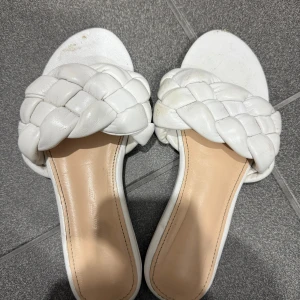 Vita flätade sandaler i skinn - Säljer ett par vita sandaler med breda, flätade remmar i mjukt skinn. De har öppen tå och platt sula, vilket gör dem både snygga och bekväma. Perfekta för sommarens alla tillfällen och ger en fräsch vibe till din outfit.