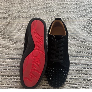 Svarta sneakers med nitar från Louboutin - Säljer ett par svarta sneakers från Louboutin i mocka med coola svarta nitar på tån och klassisk röd sula. Skorna har snörning och en platt sula, perfekt för dig som vill sticka ut med en edgy vibe.