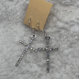 Glittriga korsörhängen i silver - Säljer ett par snygga örhängen med korsform, täckta av glittrande stenar i silverfärg. Perfekt för dig som vill ha en statement-accessoar med lite edge. Hängande modell som ger extra bling till din look.