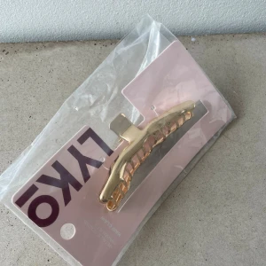 Guld hårklämma från Lyko - Snygg och minimalistisk hårklämma i metall med guldig finish från Lyko. Klämman har en stilren, böjd form och öppna sidor som ger ett trendigt uttryck. Perfekt för att snabbt fixa frisyren och addera lite extra shine till din look.