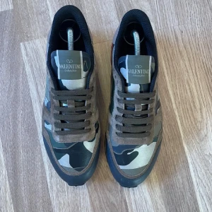Valentino Garavani Rockrunner sneakers - Säljer ett par Valentino Garavani Rockrunner sneakers med camouflagemönster i grönt, beige och svart. Skorna har detaljer i röd mocka på hälen, bruna och svarta paneler, samt platt sula med mönstrad undersida. Klassisk snörning och coola nitar på baksidan.