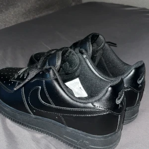 Svarta Nike Air Force 1 sneakers - Säljer ett par helsvarta Nike Air Force 1 sneakers i klassisk low-modell. Skorna har ovandel i skinn, perforerad tå och tydliga Nike-loggor på sidan och hälen. Platt sula och snörning framtill. Perfekta för dig som gillar en clean och tidlös streetstil.