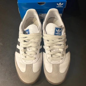 Adidas Samba OG vita sneakers - Klassiska Adidas Samba OG sneakers i vitt skinn med svarta ränder och grå mockadetaljer vid tån. Snygg retrostil med gummisula och blå Adidas-logga på plösen. Perfekta för dig som gillar tidlös streetwear och sportig vibe.