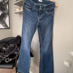 Blå bootcut jeans med broderade fickor - Snygga blå jeans från Nelly med bootcut passform och låg midja. Baktill har fickorna ett coolt vitt broderat mönster. Jeansen har råa, fransiga kanter längst ner och klassiska detaljer som dubbla knappar och fickor fram. Perfekta för dig som gillar en avslappnad stil.