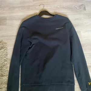 Svart sweatshirt från Lyle & Scott - Svart sweatshirt från Lyle & Scott med broderad gul logga på ärmen och diskret vit text på bröstet. Tröjan har rund hals, långa ärmar och är tillverkad i mjuk bomull. Perfekt för dig som gillar stilrena och klassiska plagg med en modern touch.