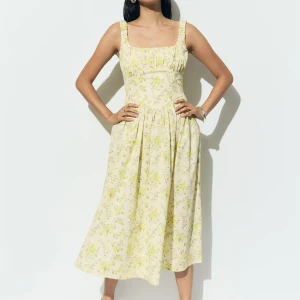 Corset Dress från GinaTricot  - Superfin långklänning från GinaTricot i ljusgul ton med blommigt mönster. Klänningen har breda axelband, smockad byst och snörning i ryggen för extra snygg passform. Tillverkad i mjuk bomull, perfekt för sommarens alla tillfällen. Använd endast en gång. Slutsåld på hemsidan.