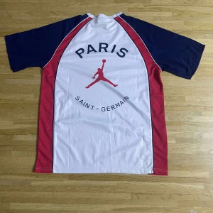PSG x Jordan vit/blå/röd fotbollströja - Snygg PSG fotbollströja i samarbete med Jordan. Tröjan är vit med marinblå och röda paneler, har korta ärmar och klassisk rund hals. PSG-logga och Jumpman-märke på bröstet, samt stor Paris Saint-Germain-tryck på ryggen. Tillverkad i lätt polyester som andas.