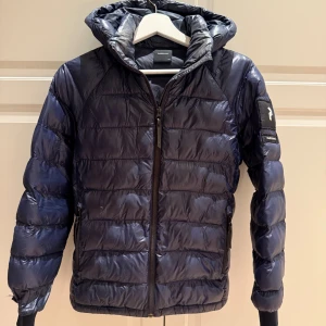 Mörkblå pufferjacka med huva - Säljer en mörkblå pufferjacka från Peak Performance med huva och dragkedja framtill. Jackan har quiltad design, två fickor med dragkedja och ribbade muddar vid ärmsluten. Tyget är glansigt. Perfekt för kyliga dagar och riktigt skön att bära. Höger ärm har en lagning som syns om man tittar nära.