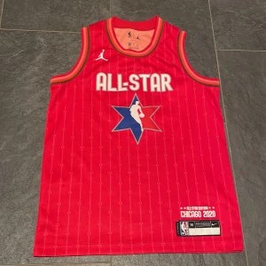 NBA All-Star Curry tröja röd M - Säljer en röd NBA All-Star Edition Chicago 2020 baskettröja med Curry #30 på ryggen. Tröjan är ärmlös, har vita detaljer och blå stjärnlogo framtill. Tillverkad i Dri-Fit polyester för bästa komfort under träning eller match.