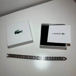 Lacoste silverfärgat armband - Stilrent armband från Lacoste i silverfärgad metall med rektangulära länkar och graverade mönster. På ena länken syns den klassiska Lacoste-krokodilen. Kommer med original ask och certifikat. Perfekt accessoar för dig som gillar sportig och modern stil.