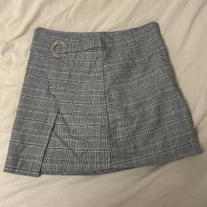 Grå rutig kortkjol med metallring - Snygg grå kortkjol med rutigt mönster och en metallring i midjan. Det står storlek 8 så tror det är en XS elr S men förmoten för M 