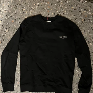 Svart sweatshirt från Les Deux - Svart sweatshirt från Les Deux med broderad vit logga på bröstet. Tröjan har rund halsringning, ribbade muddar och är långärmad. Tillverkad i mjuk bomull, perfekt för en clean och stilren look.