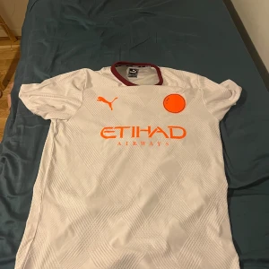 Vit Manchester City fotbollströja Puma - Säljer en vit Manchester City fotbollströja från Puma med subtilt rutmönster. Tröjan har korta ärmar, vinröd krage och orangea detaljer med Etihad Airways-logga på bröstet. Perfekt för dig som vill sticka ut på planen eller läktaren.