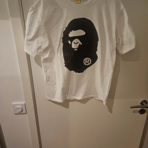 Vit BAPE t-shirt med stor ape-logga - Vit t-shirt från A Bathing Ape med stor svart ape-logga på bröstet och baksidan. Klassisk passform, rund hals och korta ärmar. Tillverkad i mjuk bomull och har en liten BAPE-tag på ärmen. Perfekt för dig som gillar streetwear och vill sticka ut.