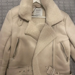 Beige teddyjacka köpt från Zalando - Säljer en beige teddyjacka från The Iconic Jacket köpt från Zalando  med bred krage och mjukt teddyfoder. Jackan har dragkedjor på fickorna, bälte i midjan och detaljer i syntetmaterial. Använt en vinter och köpt för över 1000kr. 🥰