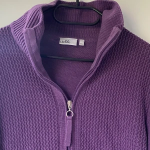 Lila stickad kofta från Isolde - Mysig lila kofta från Isolde i storlek XXL. Koftan är stickad med strukturmönster framtill, har ribbade muddar och hög krage. Stängs med dragkedja hela vägen och har långa ärmar. Perfekt för lager-på-lager under kyliga dagar.