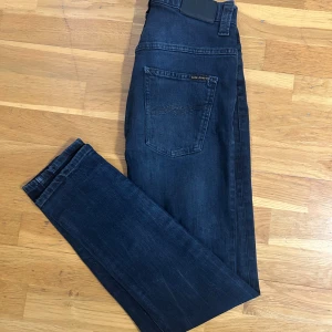 Mörkblå skinny jeans från Nudie - Snygga mörkblå jeans från Nudie med klassisk femficksdesign och diskret logga på bakfickan. Jeansen har en tight passform och raka ben, tillverkade i stretchig denim för extra komfort. Perfekta för dig som gillar en stilren och modern look.