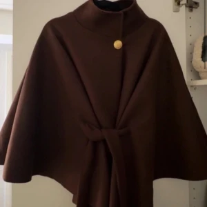 Mörkbrun kappa med bälte - Säljer en stilren mörkbrun kappa med hög krage och ett snyggt guldknapp vid halsen. Kappan har ett bälte i midjan för extra passform och en elegant, svepande siluett. Perfekt för dig som vill ha en klassisk look med modern twist.
