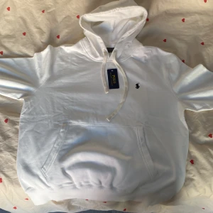 Vit hoodie från Polo Ralph Lauren - Vit hoodie från Polo Ralph Lauren med klassisk huva och snörning. Tröjan har en broderad logga på bröstet och en stor magficka framtill. Tillverkad i mjuk bomull som känns skön mot huden. Perfekt för chill dagar och enkel att matcha med jeans eller joggers. Priset är inte heller hugget i sten✌️