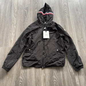 Svart vindjacka från Moncler med huva - Svart vindjacka från Moncler med huva och coola röd-vita detaljer på kanten. Jackan har dragkedja framtill, snörning i huvan och två fickor med knappar. Materialet är lätt och syntetiskt, perfekt för blåsiga dagar. Stilsäker och sportig look.