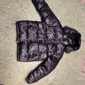 Svart pufferjacka från Polo Ralph Lauren - Snygg svart pufferjacka från Polo Ralph Lauren med glansig finish och röd broderad logga på bröstet. Jackan har huva, dragkedja framtill och flera praktiska fickor. Perfekt för kalla dagar och riktigt skön att bära.