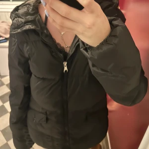 Svart dunjacka från Gant - Svart dunjacka från Gant med huva och dragkedja framtill. Jackan har två fickor med knapp, samt broderad Gant-logga på bröstet. Strl S. Skada vid dragkedjan inget som syns när den är stängd!💓