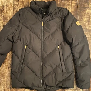 Svart pufferjacka från Reima, 13 år - Svart pufferjacka från Reima med snyggt quiltat mönster och hög krage. Jackan har dragkedja framtill, två fickor med dragkedja och Reima-logga på ärmen. Perfekt för kalla dagar och riktigt skön att bära.