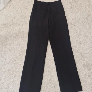 Svarta kostymbyxor från Lindex - Klassiska svarta kostymbyxor från Lindex med rak passform och hög midja. Byxorna har hällor för bälte och stängs med knapp och dragkedja. Perfekta för en clean och stilren look.