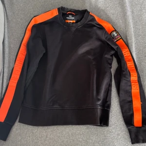 Svart fleece-tröja Parajumpers YS - Svart fleece-tröja från Parajumpers med orangea ränder längs ärmarna och broderad logga på axeln. Tröjan har rund hals, ribbade muddar och texten 'PARAJUMPERS' på ärmarna. Perfekt för dig som gillar sportig och cool stil.