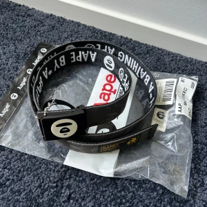 AAPE reversible bälte - Snyggt bälte från Aape by A Bathing Ape med tydlig logga på metallspännet och text längs hela bandet eller camo. Bältet har en urban streetwear-vibe och är gjort i slitstark syntet. Perfekt accessoar för att lyfta din outfit. Pris kan diskuteras.