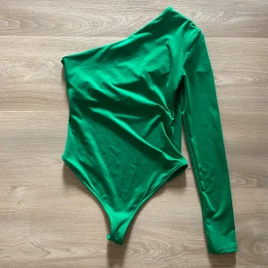 Grön oneshoulder body från Zara - Säljer en grön body från Zara i storlek S. Den har en snygg oneshoulder-design med lång ärm och knäppning i grenen. Tillverkad i mjukt och stretchigt material som sitter tight och bekvämt. Perfekt för att skapa en cool och trendig look.