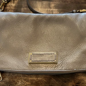 Gråbeige axelväska från Marc by Marc Jacobs - Snygg beigegrå axelväska i mjukt skinn från Marc by Marc Jacobs. Väskan har justerbar rem, guldfärgade metalldetaljer och flera fack med dragkedja. Perfekt storlek för det viktigaste och stilren design med logoplatta framtill.
