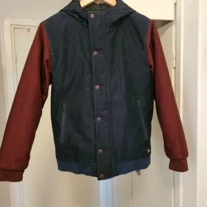 Vans jacka mörkblå/vinröd obs! youth size L - Snygg jacka från Vans i storlek L i ungdomsstorlek. Marinblå och vinröd. Jackan har huva, dragkedjefickor och dragkedja och tryckknappar framtill. Mått hängande på galgen, så blir inte exakt på millimetern. Från slutet på axeln till längst ner på mudden: 57 cm. Längd/Nacksömmen till längst ner: 64 cm. Armhåla till armhåla rakt över (lite utsträckt): 52 cm.