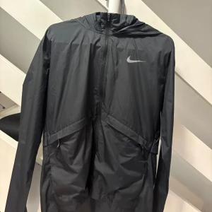 Nike Miller jacka Svart - Svart vindjacka från Nike med huva och hel dragkedja framtill. Jackan har ett stilrent Nike Swoosh-tryck på bröstet och meshfoder på insidan för extra ventilation. Perfekt för löpning eller andra utomhusaktiviteter. Snygg och funktionell design.