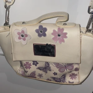 Vintage Minnie Mouse-väska med fjärilar och blommor - Ljus axelväska med lila fjärilar och blommor, samt metallmärke med Minnie Mouse-logga framtill. Justerbar axelrem och små handtag – kan bäras både på axeln eller i handen. Ger en riktig 2000-tals/Y2K-look!