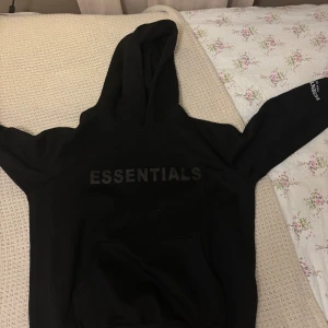Svart Essentials Fear of God hoodie - Svart hoodie från Essentials Fear of God med stor logga på bröstet och mindre tryck på ärmen. Klassisk känguruficka framtill och huva. Använd två gånger ungefär, kände att den inte passa mig helt!