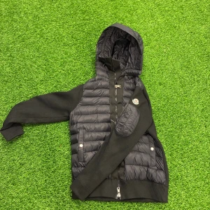 Svart moncler cardigan  - Svart pufferjacka från Moncler med stickade ärmar och quiltad kropp. Jackan har huva, dragkedja framtill, två fickor med tryckknappar och en patch med Moncler-logga på ärmen. Materialmix av polyester och stickat ger en modern look.