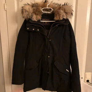 Svart parkas från Woolrich med pälskrage - ❤️Modellen ”Valetine”❤️ Säljer en svart parkas från Woolrich med stor fluffig pälskant på huvan. Jackan har dragkedja och knappar framtill, två stora fickor och en klassisk längre passform. Perfekt för kalla dagar med sin varma och stilrena look. 