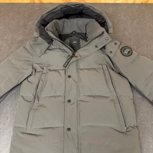 Grå dunjacka från Canada Goose M - Säljer en grå dunjacka från Canada Goose i storlek M. Jackan har huva, hög krage, dragkedja och tryckknappar framtill samt två stora fickor. Klassisk patch på ärmen och stilren design som håller dig varm under kalla dagar. Päls kommer med i köpet! Samt tags och liknande även canada goose påsen