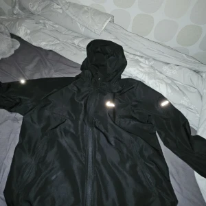 Svart vindjacka från Nike med huva - Svart vindjacka från Nike med huva och dragkedja framtill. Jackan har två sidofickor med dragkedja och en stilren Nike-logga på bröstet. Perfekt för blåsiga dagar och sportiga outfits.