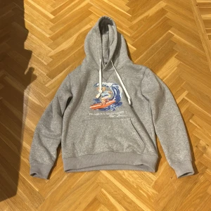 Grå hoodie Polo Ralph Lauren björn  - Grå hoodie från Polo Ralph Lauren med surfande björn-tryck på bröstet. Tröjan har huva med vita snören, magficka och ribbade muddar. Perfekt för dig som gillar streetstyle och vill ha något bekvämt och snyggt.