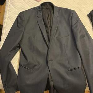 Svart kavaj från Bläck - Stilren svart kavaj från Bläck med klassisk passform och snygga detaljer. Kavajen har två framfickor, bröstficka och slät rygg. Fodret är svart med Bläck-logga och insidan har en vit kant. Perfekt för dig som vill ha en clean och modern look.