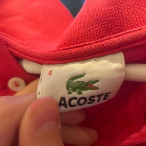 Röd kortärmad skjorta från Lacoste - Riktigt snygg röd skjorta från Lacoste med klassisk krokodillogga på bröstet. Skjortan har korta ärmar, krage och knappar framtill. Materialet är mjuk bomull som känns skönt mot huden. Perfekt för dig som gillar stilrena och sportiga plagg.