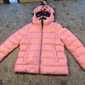 Rosa skidjacka AI Riders stl 16 år - Säljer en rosa skidjacka från AI med coola inbyggda svarta goggles i huvan. Jackan har dragkedja framtill, två fickor och quiltad design. Storlek 16 år.
