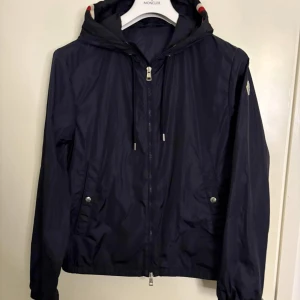 Mörkblå vindjacka från Moncler - Snygg mörkblå vindjacka från Moncler med huva och dragkedja framtill. Jackan har Moncler-logga på ärmen, diskreta fickor med tryckknappar och röda/vita detaljer på huvan. Perfekt för blåsiga dagar och stilren look. Självklart är jackan äkta!!! Tveka inte att skriva om ni har funderingar😁