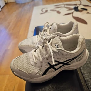 Vita Asics sneakers med mesh och snörning - Säljer ett par fräscha vita Asics sneakers i storlek 33. Skorna har ventilerande mesh på ovandelen, klassisk snörning och svarta Asics-loggor på sidorna. Perfekta för dig som vill ha bekväma och snygga skor till vardagen eller träningen.