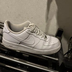 Nike Air Force 1 vita sneakers - Fräscha Nike Air Force 1 sneakers i helvitt med klassisk låg siluett. Skorna har snörning, perforerad tå och rundad form. Tillverkade i skinn med vit sula och ikonisk Swoosh på sidan. Perfekta för dig som gillar clean och tidlös streetstil.