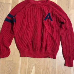 Röd stickad tröja Iconic Sport S - Röd stickad tröja från Iconic Sport med blåa ränder på ena ärmen och ett blått A på bröstet. Tröjan har grovstickad struktur, ribbade muddar och rund hals. Perfekt för dig som gillar sportig och retro stil.