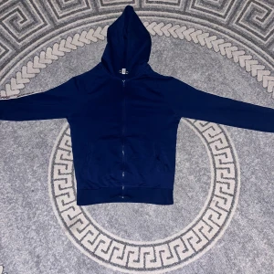 Marinblå Moschino hoodie med dragkedja - Säljer en marinblå hoodie från Moschino med hel dragkedja och huva. Tröjan har långa ärmar och vita band med Moschino-logga längs ärmarna. Tillverkad i mjuk bomull, perfekt för chill dagar. Snygg och stilren design med ribbade muddar.